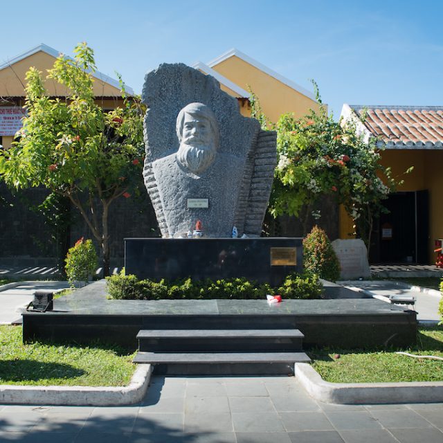 Un monument en pierre représentant un buste en bas-relief de Kazimierz Kwiatkowski, posé sur un socle sombre, dans une petite place de Hoi An, avec des arbres et des bâtiments jaunes en arrière-plan.