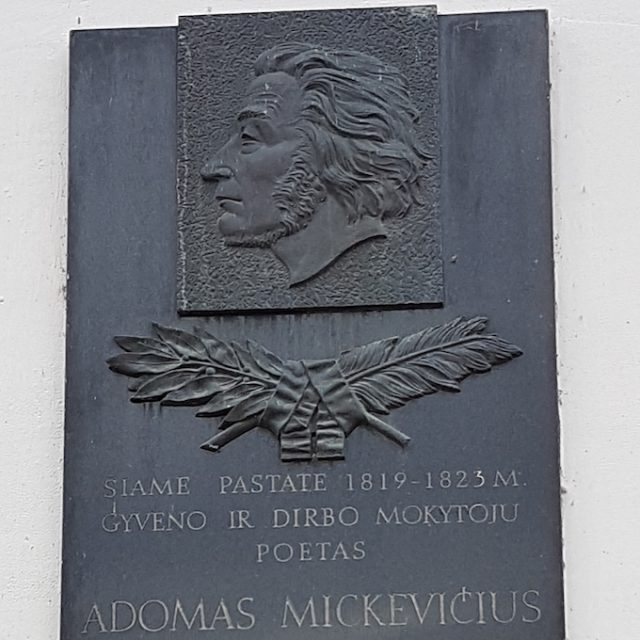 Tablica pamiątkowa w Kownie z reliefowym profilem Adama Mickiewicza, skrzyżowanym piórem i gałązką laurową oraz litewskim napisem na ścianie budynku.