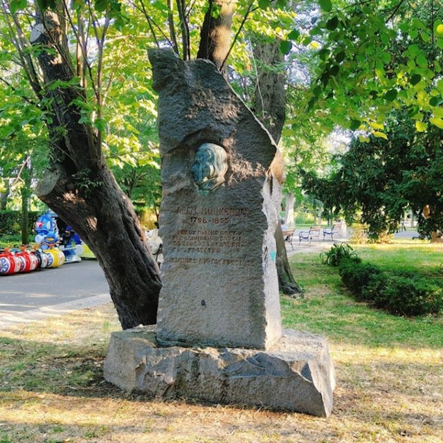 Stone monument to Adam Mickiewicz in Burgas. Rough monolith on a wide base with a bronze profile relief and a carved inscription on the front. Trees and a park path surround it.