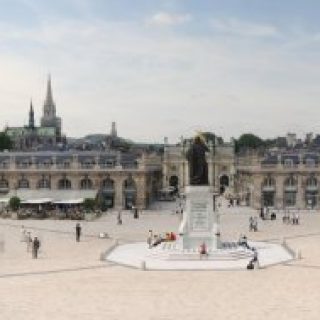 Vue panoramique de la Place Stanislas à Nancy avec des bâtiments classiques symétriques, une statue centrale et des passants. La place est entourée de façades ornées sous un ciel dégagé.